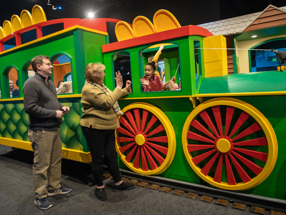 Two adults standing alongside the green Dinosaur Train replica and waving a at a child whose head is sticking out the window. and waving back